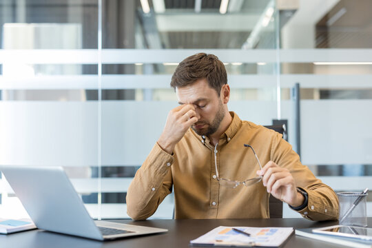 Stressed businessman feeling tired and suffering from a headache, rubbing his nose bridge while holding eyeglasses, experiencing burnout from work at his modern office desk
