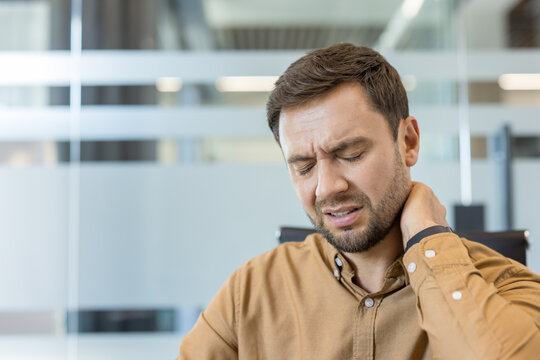 Caucasian man experiencing severe neck pain and discomfort, holding his hand to the back of his neck, representing stress, muscle tension, and health issues in an office environment