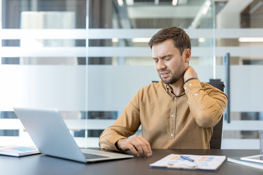 Young man sitting at his desk in a modern office, experiencing severe neck pain and discomfort while working on a laptop, indicating stress and fatigue from prolonged computer use