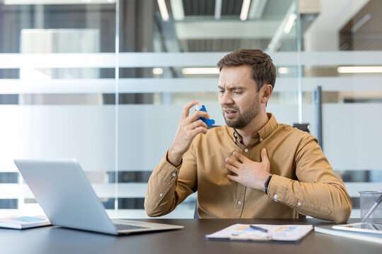 Young man using a blue medical inhaler for asthma, experiencing breathlessness and chest pain while sitting at his desk in a modern office, working on a laptop