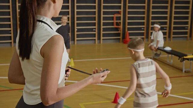 Caucasian female coach holding stopwatch and timing children practicing soccer drills in school gymnasium. Footage ideal for youth sports and school curriculum marketing