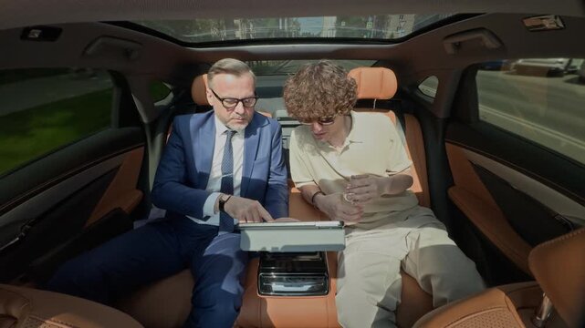 Middle aged Caucasian businessman showing tablet to young Caucasian man drinking water in backseat of moving luxury car. Video ideal for wealth management or mentorship ads
