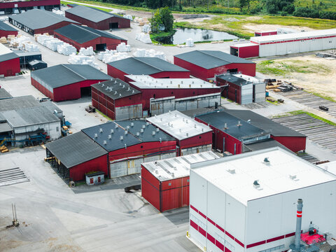 Aerial view of a large timber production facility with red industrial buildings and stacks of sawn timber in the rural landscape of Lauciene Parish, Talsi Municipality, Latvia.
