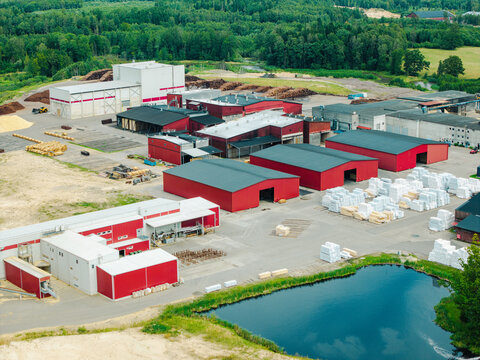 Aerial view of a wood processing facility with red industrial buildings, stacks of lumber, and a small pond in Lauciene Parish, Talsi Municipality, Latvia.