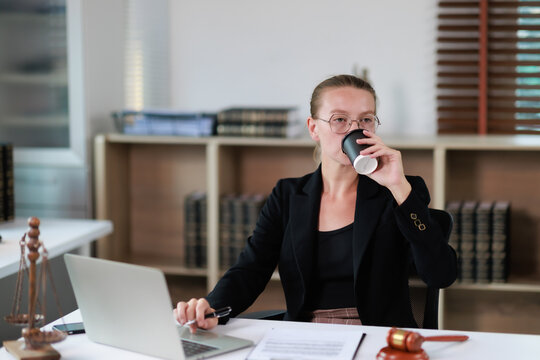 Female lawyer in a formal suit and eyeglasses drinking coffee from a disposable cup while working on her laptop in a professional law office setting