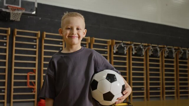 Portrait of Caucasian child boy holding soccer ball in school gym, smiling at camera. Footage is perfect for promoting youth sports programs, physical education, or active lifestyle campaigns
