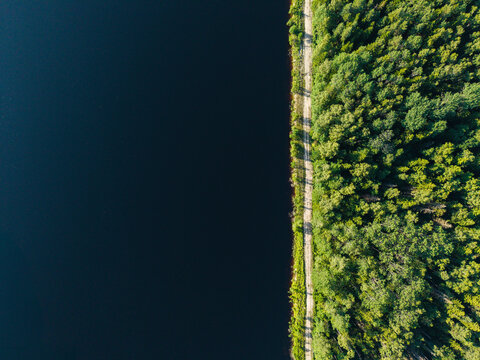 Aerial view of a straight dirt path separating a dark body of water from a dense green forest at Kalnciema dolomitu karjers in Valgunde Parish, Jelgava Municipality, Latvia.