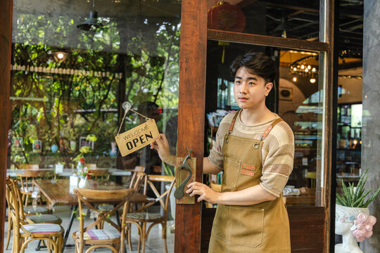 Young barista cafe owner startup smiling at entrance with open sign visible, confident entrepreneur representing hospitality service and customer readiness, positive mood in small business operation.