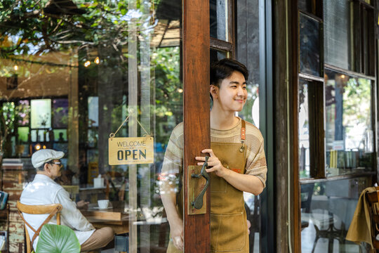 Yong small business owner opening cafe door with open sign displayed, inviting customers inside cozy coffee shop and restaurant, daily startup routine reflecting growth and opportunity in business.