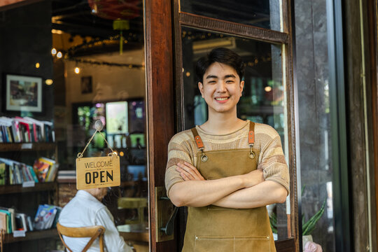 Young barista cafe owner smiling at entrance with open sign visible, confident entrepreneur representing hospitality service and customer readiness, positive mood in small business operation.