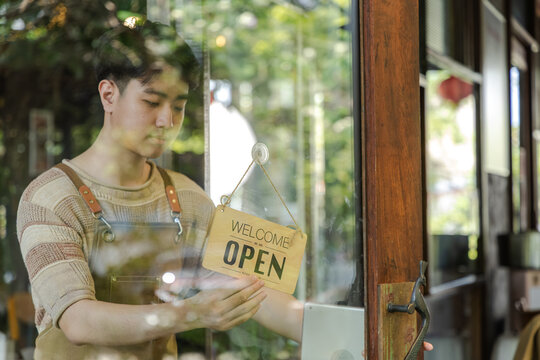 Small business startup, young cafe owner barista looking outside through door with closed sign reflecting concern about business situation and customer flow in coffee shop, worker flipped open sign
