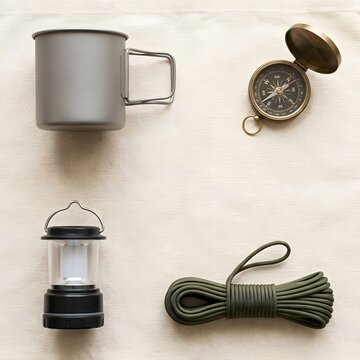 Flat lay of minimalist camping gear with a titanium mug, brass compass, LED lamp and paracord on canvas