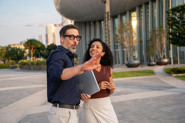 Fototapeta premium Two diverse business professionals standing outside a modern glass office building, looking at a discussing a project during a meeting. 