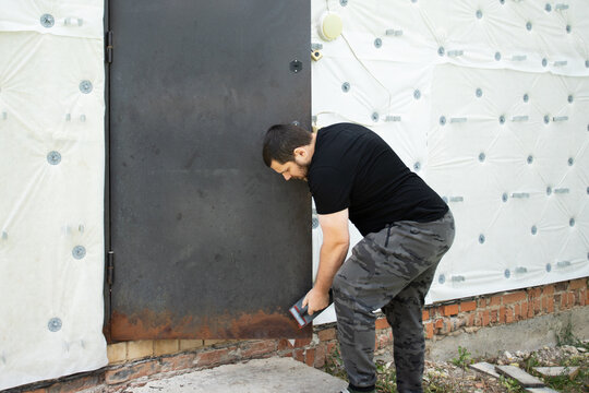 male homeowner sanding severe corrosion from an aging exterior steel door, carrying out diy surface preparation before repainting and residential property repair
