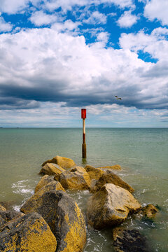 Red navigation marker beyond a rocky groyne under dramatic clouds at Waxham Beach near Horsey Gap on the North Sea coast of Norfolk, England.