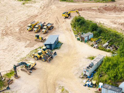 Aerial view of a gravel pit with yellow excavators, dump trucks, and construction equipment parked on sandy ground in Slampe parish, Tukums Municipality, Latvia.