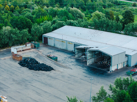 Aerial view of a waste management service facility with piles of discarded tires, industrial dumpsters, and a large warehouse building surrounded by lush green trees in Jurmala, Latvia.