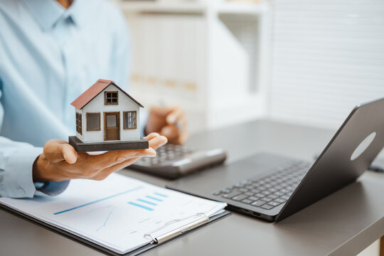 Home model is presented above mortgage paperwork near a laptop and calculator, linking property insurance, finance review, and residential. Realestate property, mortgage; realestate focus.