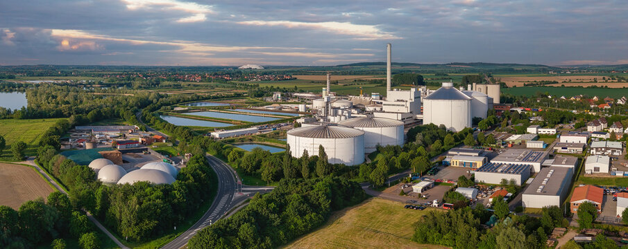 Aerial view of the industrial sugar factory facility featuring large white storage tanks, a tall chimney, and surrounding green fields under a soft sunset sky.