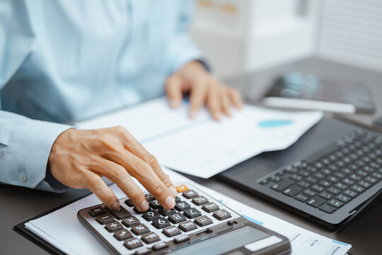 Business hand uses a calculator over paperwork beside a laptop, showing financial analysis, numerical data review, and accounting report work. Using calculator, budget; using focus.