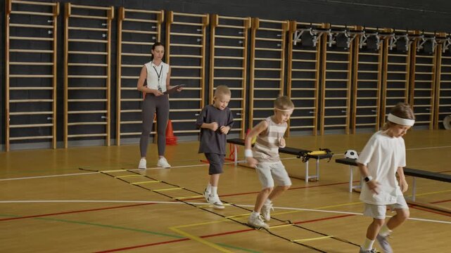Caucasian boys practicing agility ladder drills in school gymnasium while female Caucasian coach watching with clipboard. Footage is ideal for promoting youth sports programs and physical education