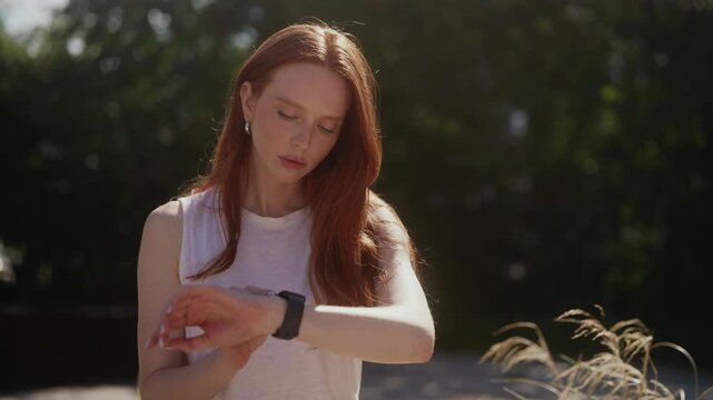 Close-up portrait of young beautiful redhead sporty woman with freckles setting up smartwatch and smiling, checking fitness results after successful outdoor training session on sunny day in park.