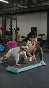 Vertical video of Caucasian young adult female trainer timing Caucasian male child holding plank on step platform in gym. Footage is ideal for youth fitness coaching promos