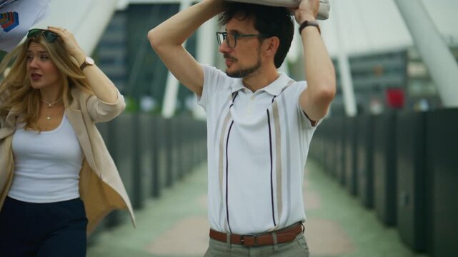 Two colleagues are caught unprepared in a sudden rain shower on a city bridge. They use their bags and documents to shield themselves from the weather.