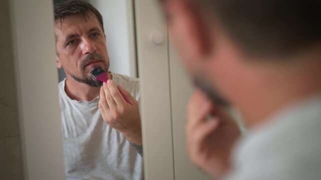 Middle-aged man carefully applying black beard dye to graying goatee with pink brush while looking in bathroom mirror, covering gray facial hair during home grooming routine.