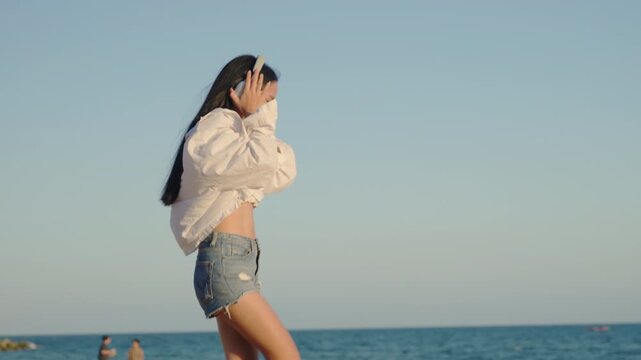 Young woman enjoying music and dancing on beach wearing wireless headphones, beautiful long haired girl in white shirt and denim shorts listening to playlist by sea under clear sky.