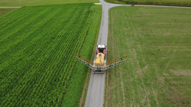 Aerial view of a red tractor pulling a yellow slurry tanker along a narrow farm road between a maize field and a meadow, while the trailing-hose applicator boom unfolds to working position