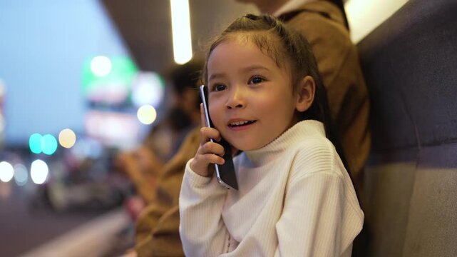 child girl talking on a smartphone with her mother while sitting on a bench at a bus station in city at night
