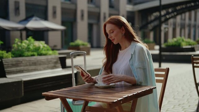 Beautiful redhead traveler with luggage sitting at table in sunlit outdoor cafe, leisurely drinking coffee and scrolling through smartphone waiting for flight at airport terminal or train station.