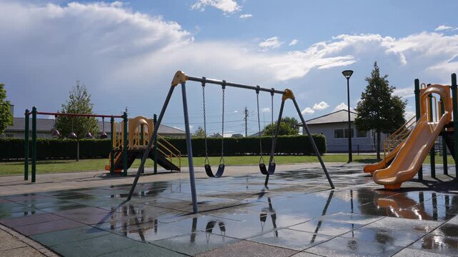Empty swings at a wet children's playground moving in the wind after a summer rain shower, reflecting the cloudy sky on the wet ground in a public park