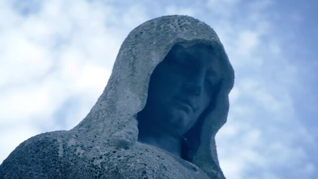 Weathered hooded stone statue in a cemetery against a cloudy blue sky. The camera slowly rises, revealing the textured details of the ancient monument. Evokes themes of remembrance and timelessness.
