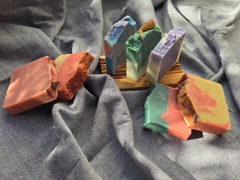 Vibrant, multicolored handmade cold process soap bars topped with dried rose buds and citrus slices, featuring eco-friendly corrugated cardboard packaging on a rustic grey fabric background.