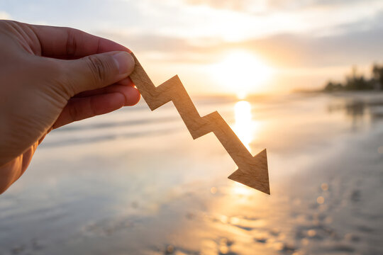 Hand holding wooden downward trend graph against sunset beach backdrop symbolizing economic decline