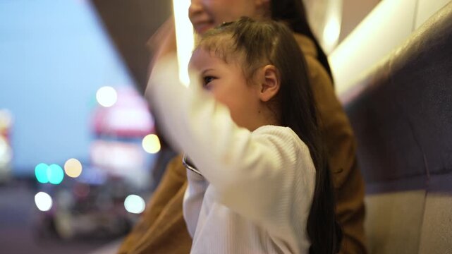 child girl talking on a smartphone with her mother while sitting on a bench at a bus station in city at night