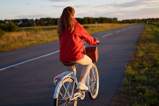 A person rides a bicycle along a quiet road in the countryside. The sun sets in the background, and fields stretch out on either side of the path