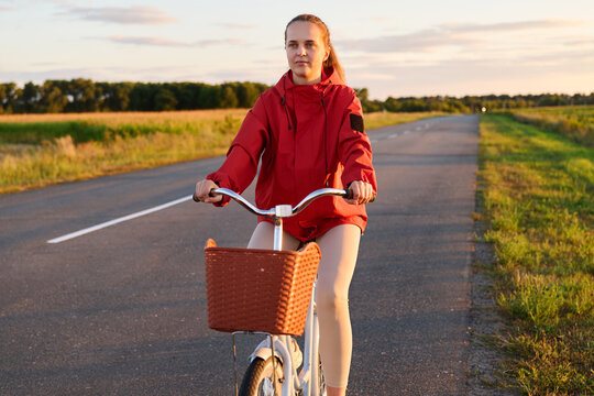 A person rides a bicycle on a road surrounded by fields in a rural area. The sun is setting, and the sky shows warm colors as the day ends