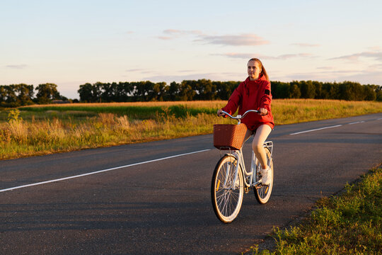 A person cycles on a road lined with fields. The sun sets in the background, casting warm light on the scene. The cyclist enjoys the ride in a casual outfit