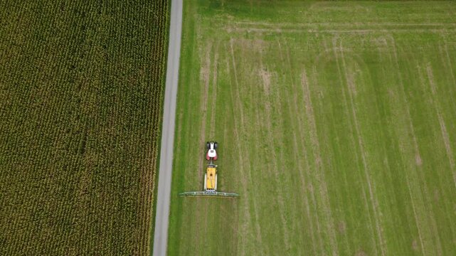 High-altitude top-down aerial view of a red tractor pulling a yellow slurry tanker as it spreads liquid manure across a meadow with a trailing-hose applicator