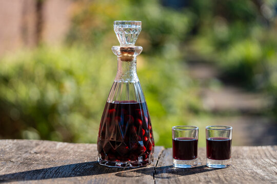 Crystal decanter filled with red cherry liqueur and whole berries, next to two filled shot glasses on rustic wooden table outdoor, closeup. Red fruit alcohol drink or traditional tincture
