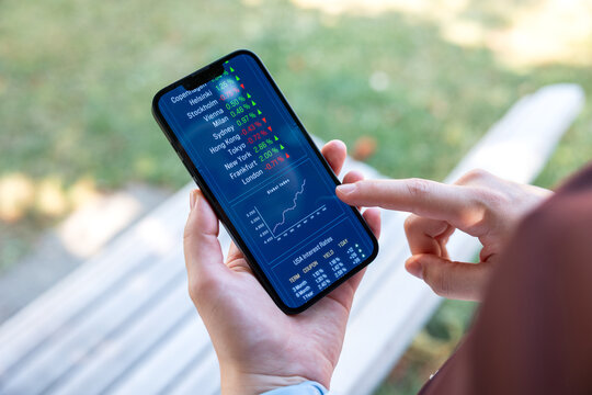 Woman behind a bench points on a smart phone with stock maket data, chart. Trading, stockbroker, investment decision, stock prices, financial advisor, assets, wealth.