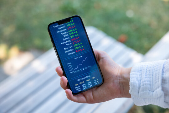 Woman behind a bench, holding a smart phone with stock maket information, chart and market data. Stockbroker, investment decision, stock prices, financial advisor, assets, wealth.