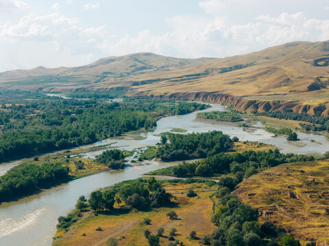 Aerial view of the winding river flowing through the lush green valley with rolling hills and golden fields under a bright sky in Mtskheta, Mtskheta-Mtianeti, Georgia.