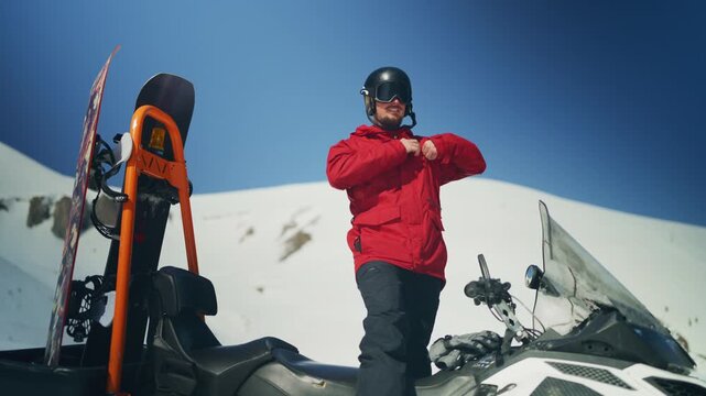 A male snowboarder in a red jacket adjusts his gear next to a snowmobile with skis and a snowboard. He is preparing for winter sports in a sunny mountain environment.