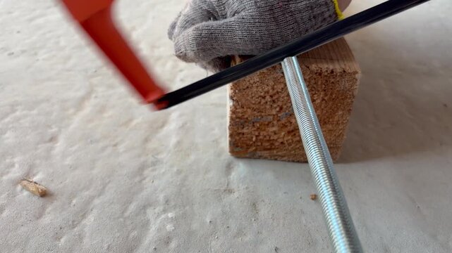 A worker cuts a threaded metal rod with a hacksaw on a block of wood. Manual labor, metalworking, DIY projects, and construction site activities involving hardware preparation.