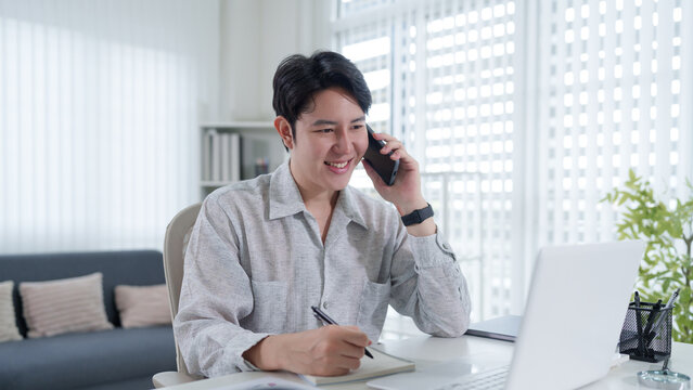 Young Asian businessman smiling while taking phone call at laptop in bright modern home office with copy space