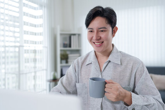 Young Asian businessman holding coffee mug smiling at camera in bright home office during hybrid work